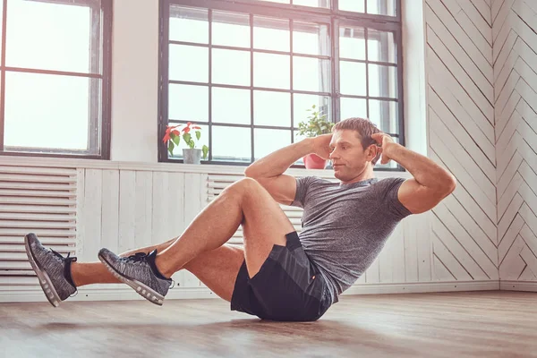 Stilig fitness man i en t-shirt och shorts gör magövningar på golvet hemma. — Stockfoto