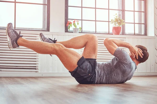 Hombre de fitness guapo en una camiseta y pantalones cortos haciendo ejercicios abdominales en el suelo en casa . — Foto de Stock