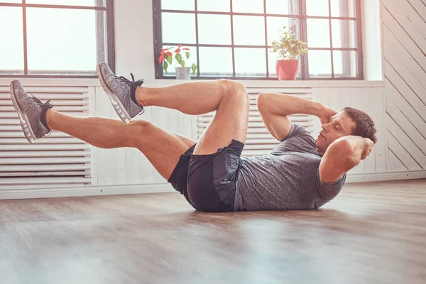 Stilig fitness man i en t-shirt och shorts gör magövningar på golvet hemma. — Stockfoto