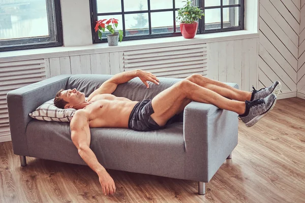 Pohledný shirtless svalnatý muž leží na gauči, odpočívá po náročném tréninku doma. — Stock fotografie