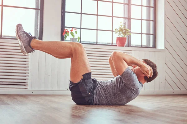 Handsome fitness man in a t-shirt and shorts doing abdominal exercises on floor at home. — Stock Photo, Image