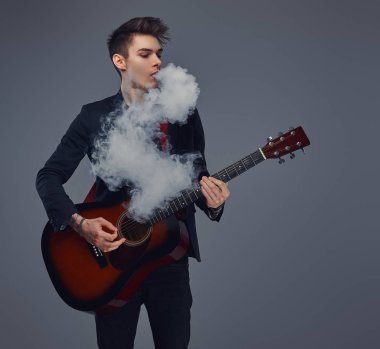 Şık giysileriyle exhales akustik gitar çalarken duman içinde şık saçlı yakışıklı genç müzisyen.