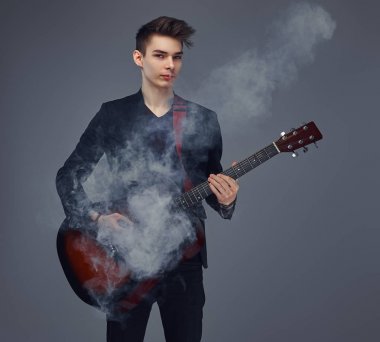 Şık giysileriyle exhales akustik gitar çalarken duman içinde şık saçlı yakışıklı genç müzisyen.