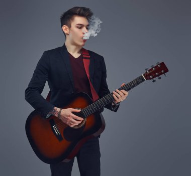 Şık giysileriyle exhales akustik gitar çalarken duman içinde şık saçlı yakışıklı genç müzisyen.