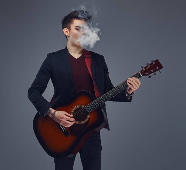 Şık giysileriyle exhales akustik gitar çalarken duman içinde şık saçlı yakışıklı genç müzisyen.