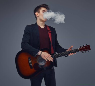 Şık giysileriyle exhales akustik gitar çalarken duman içinde şık saçlı yakışıklı genç müzisyen.