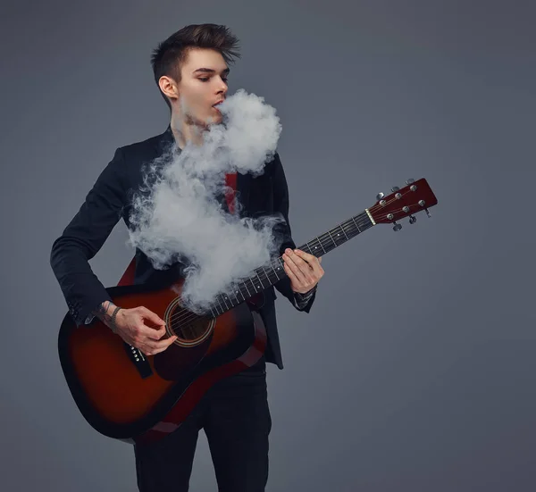 Handsome young musician with stylish hair in elegant clothes exhales smoke while playing acoustic guitar. — Stock Photo, Image