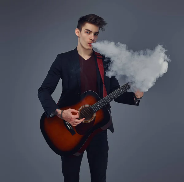 Guapo joven músico con el pelo elegante en ropa elegante exhala humo mientras toca la guitarra acústica . — Foto de Stock