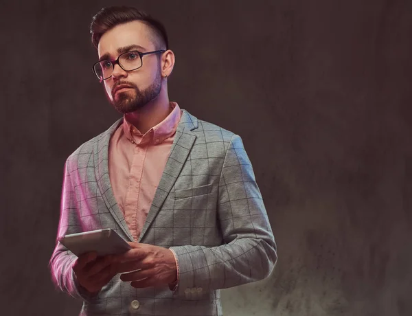 Nahaufnahme Porträt eines selbstbewussten bärtigen Mannes mit Frisur und Sonnenbrille im grauen Anzug und rosa Hemd, hält ein Tablet in einem Atelier. — Stockfoto