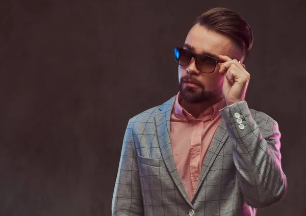 Retrato de cerca de un hombre barbudo con estilo y confianza con peinado y gafas de sol en un traje gris y camisa rosa, posando en un estudio . — Foto de Stock