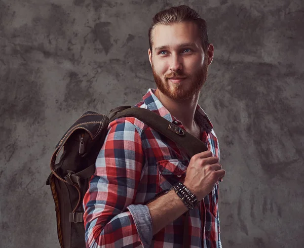 Handsome stylish redhead traveler in a flannel shirt with a backpack, posing in a studio on a gray background.