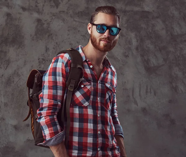 Handsome stylish redhead traveler in a flannel shirt and sunglasses with a backpack, posing in a studio on a gray background.