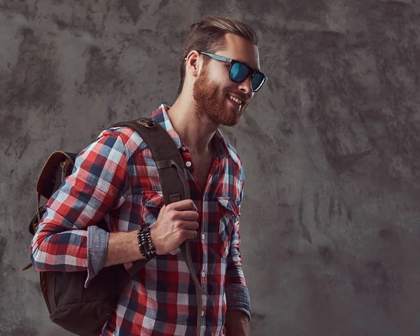 Bellissimo viaggiatore rossa elegante in una camicia di flanella e occhiali da sole con uno zaino, in posa in uno studio su uno sfondo grigio . — Foto Stock