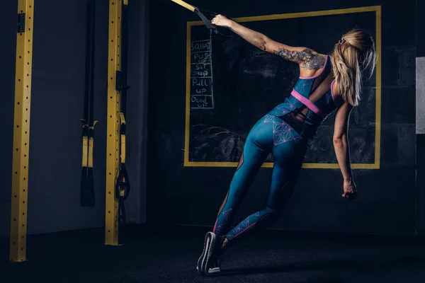 Schöne muskulöse blonde Frau in Sportbekleidung mit Tätowierung auf dem Arm beim Training mit dem Trx-System im Fitnessstudio. — Stockfoto