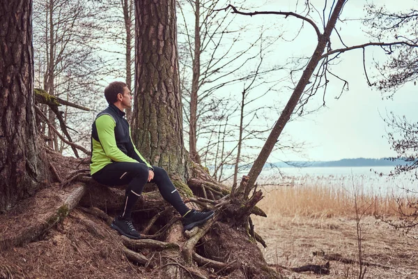 Jogger siedzi pod drzewa w lesie i wygląda od odpoczynku po dłuższej perspektywie. Aktywny styl życia. — Zdjęcie stockowe