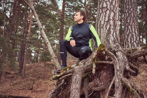 Jogger est assis sous un arbre dans la forêt et regarde loin, se reposant après une longue course. Mode de vie actif . — Photo