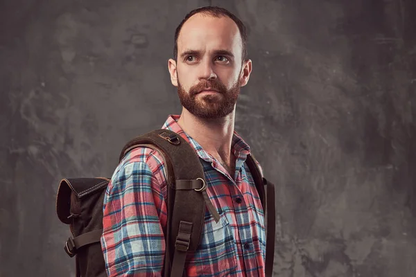 Portrait of a handsome tattooed traveler in a flannel shirt with a backpack, standing in a studio.