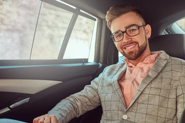 Successful stylish young businessman in a gray suit and pink shirt, riding on a back seat in a luxury car. — Stock Photo, Image