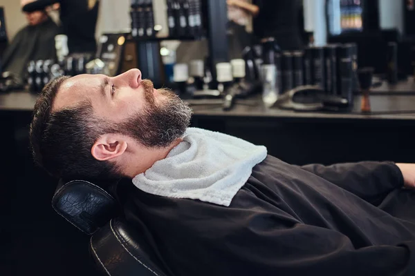 Handsome bearded male waits for the hairdresser sitting on a barber chair. — Stock Photo, Image