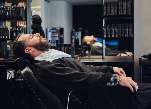 Красивий бородатий чоловік чекає на перукаря, який сидить на перукарському стільці . — стокове фото