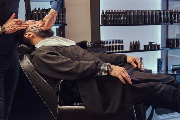 Bearded male sitting in an armchair in a barber shop while hairdresser shaves his beard with a dangerous razor. — Stock Photo, Image