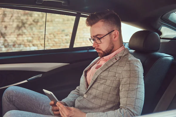 Jovem empresário bem sucedido e elegante em um terno cinza e camisa rosa, montando em um banco de trás em um carro de luxo . — Fotografia de Stock