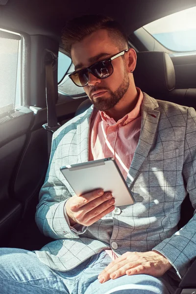 Erfolgreicher stylischer junger Geschäftsmann im grauen Anzug und rosa Hemd, der in einem Luxusauto auf der Rückbank sitzt. — Stockfoto