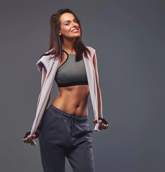 Sorridente donna bruna fitness in un abbigliamento sportivo grigio in posa in uno studio . — Foto Stock