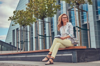 Bir gökdelen karşı parkta bir bankta oturmuş bir kitapla eğitim modern giysiler, çekici sarışın kadın.