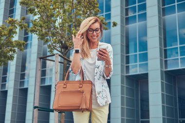 Şık giysiler ve gözlük bir çanta ile moda kadın blogger tutan bir gökdelen karşı duran bir akıllı telefon.
