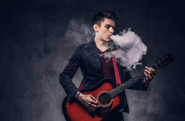 Knappe jonge muzikant met stijlvolle haren in elegante kleding uitademt rook tijdens het spelen van akoestische gitaar. — Stockfoto