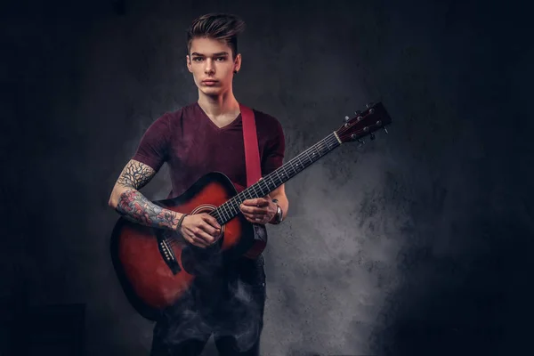 Handsome young musician with stylish hair in a t-shirt, holds a guitar in his hands and playing.