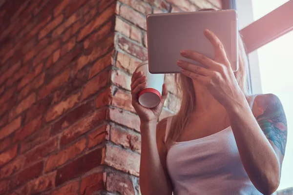 Potret seorang wanita hipster pirang memegang secangkir kopi sambil mencari sesuatu pada tablet digital di sebuah ruangan dengan interior loteng . — Stok Foto