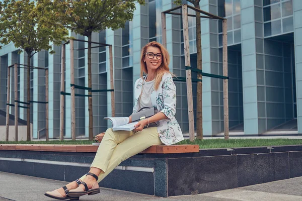 Очаровательная блондинка в современной одежде, учится с книгой, сидит на скамейке в парке против небоскреба . — стоковое фото