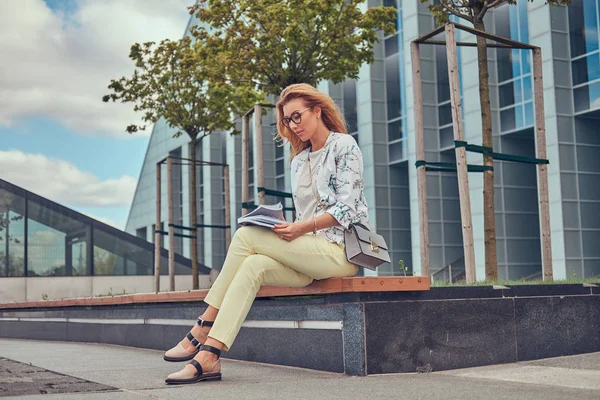 Очаровательная блондинка в современной одежде, учится с книгой, сидит на скамейке в парке против небоскреба . — стоковое фото