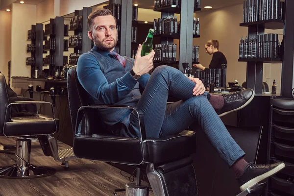 Un coiffeur professionnel assis sur un fauteuil, se repose après le travail avec une bouteille de bière à la main . — Photo