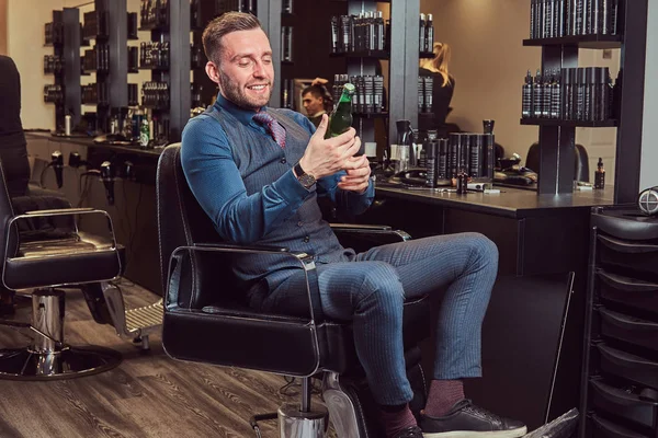 Ein professioneller Friseur sitzt auf einem Sessel und ruht sich nach der Arbeit mit einer Bierflasche in der Hand aus. — Stockfoto