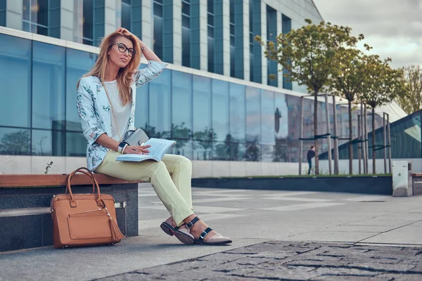 Uroczy Kobieta blonde w nowoczesne ubrania, studia z książką, siedząc na ławce w parku przed drapacz chmur. — Zdjęcie stockowe