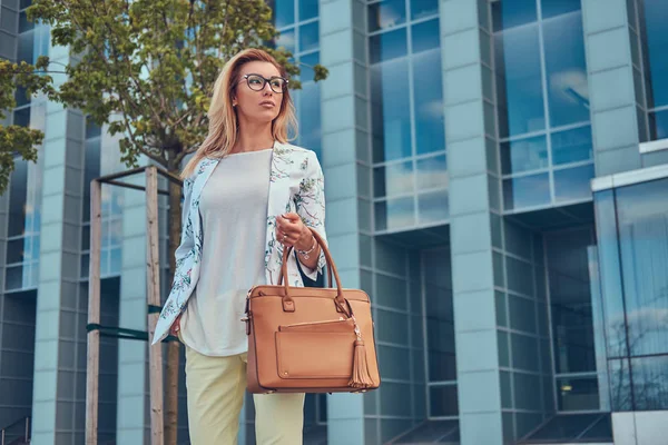 Krásná módní žena elegantní oblečení a brýle s kabelku, stojící proti mrakodrap. — Stock fotografie