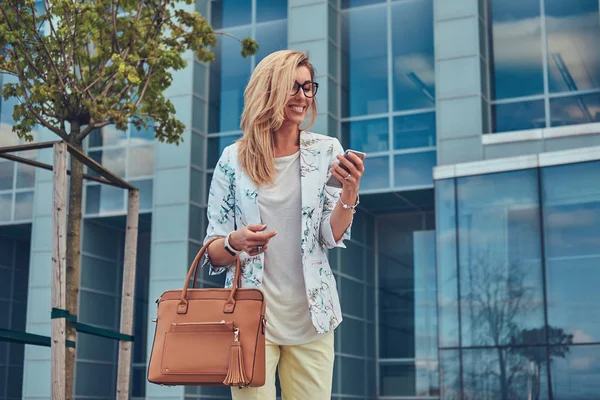 Modische Bloggerin in stylischer Kleidung und Brille mit Handtasche, hält ein Smartphone vor einem Wolkenkratzer. — Stockfoto