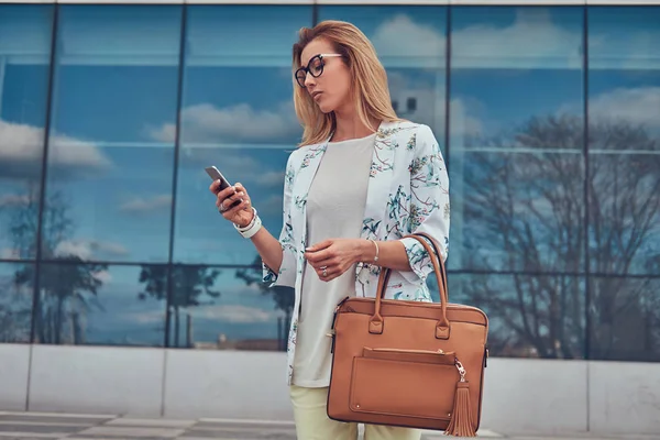 Blogger femenina de moda en ropa elegante y gafas con un bolso, sostiene un teléfono inteligente, de pie contra un rascacielos . — Foto de Stock