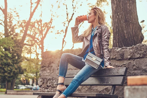 Trendige blonde Frau entspannt im Freien und trinkt Kaffee zum Mitnehmen, während sie auf der Bank im Stadtpark sitzt. — Stockfoto