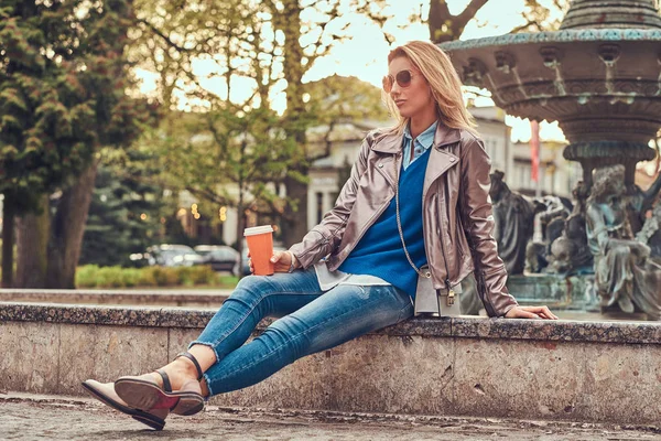 Donna bionda alla moda si rilassa all'aperto, bevendo caffè da asporto mentre si siede sulla panchina nel parco cittadino . — Foto Stock