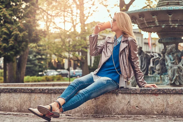 Μοντέρνο ξανθιά γυναίκα χαλαρώνει υπαίθρια, πίνοντας takeaway καφέ ενώ κάθεται στον πάγκο στο πάρκο της πόλης. — Φωτογραφία Αρχείου