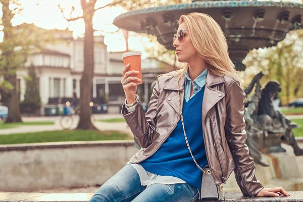 Mulher loira na moda relaxa ao ar livre, bebendo café takeaway enquanto sentado no banco no parque da cidade . — Fotografia de Stock