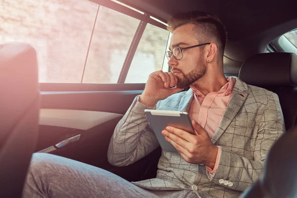 Jovem empresário bem sucedido e elegante em um terno cinza e camisa rosa, montando em um banco de trás em um carro de luxo . — Fotografia de Stock