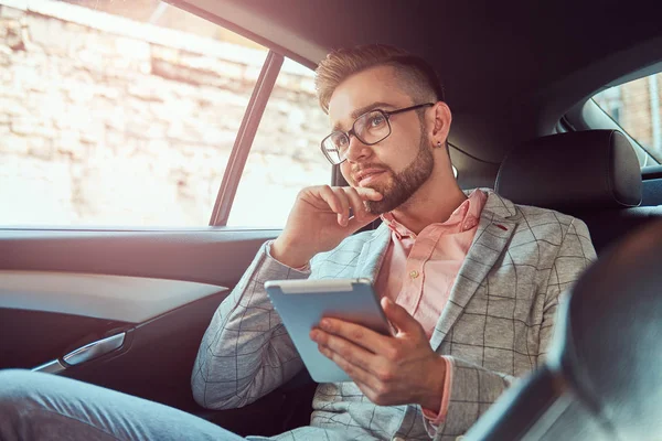 Successful stylish young businessman in a gray suit and pink shirt, riding on a back seat in a luxury car. — Stock Photo, Image