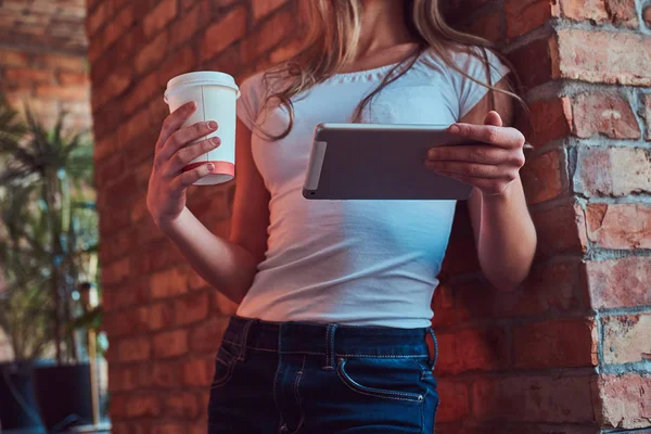 Πορτρέτο του μια νεαρή ξανθιά γυναίκα κρατά ένα φλιτζάνι takeaway καφέ και χρησιμοποιώντας ένα ψηφιακό tablet ενώ κλίνει ενάντια σε έναν τοίχο από τούβλα σε ένα δωμάτιο με εσωτερικό πατάρι. — Φωτογραφία Αρχείου
