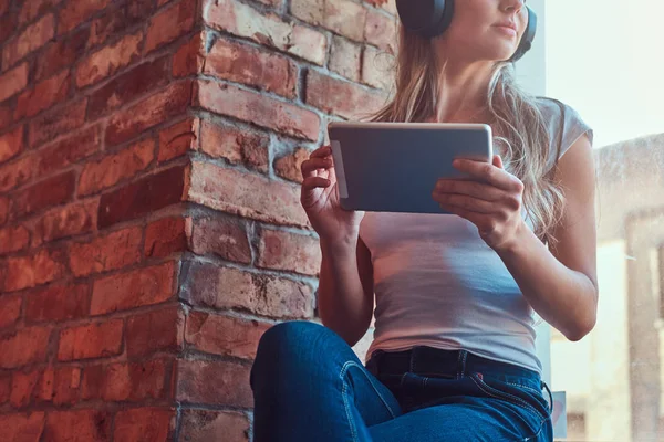 Jeune femme blonde écoutant de la musique et tenant une tablette numérique assise sur un rebord de fenêtre dans une pièce avec mezzanine intérieure . — Photo