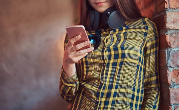 Jeune étudiante brune en tenue décontractée écoutant de la musique tout en utilisant un smartphone tout en s'appuyant contre un mur de briques dans une chambre avec mezzanine intérieure . — Photo